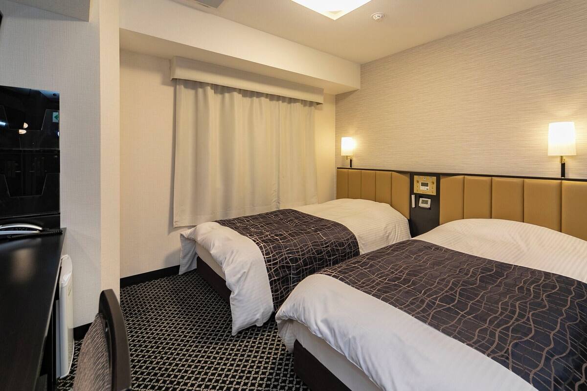 Hotellihuone, jossa on kaksi sänkyä, sängynpääty, jossa on ohjauspaneeli, ikkuna verhoilla ja kuvioitu matto.