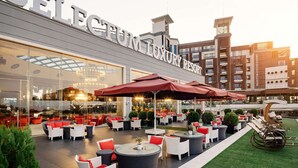 An outdoor seating area with tables and chairs, a glass facade, and a building with "SELECTION LUXURY RESORT" signage. - Selectum Luxury Resort (Serik)