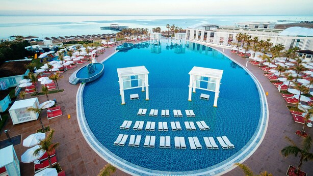 A large, circular swimming pool with lounge chairs and umbrellas, surrounded by palm trees and a view of the sea. - Selectum Luxury Resort (Serik)