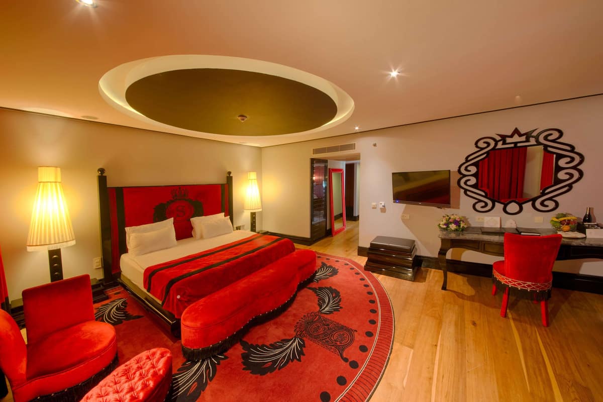 Una habitación de hotel moderna con una cama grande, sillas tapizadas en rojo, un escritorio con espejo y una alfombra redonda.