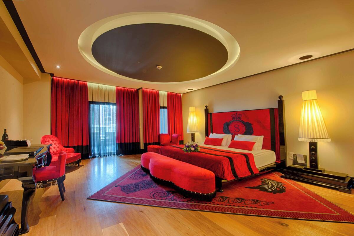 Habitación de hotel con cama, sofá rojo, piano y un ventanal con cortinas.
