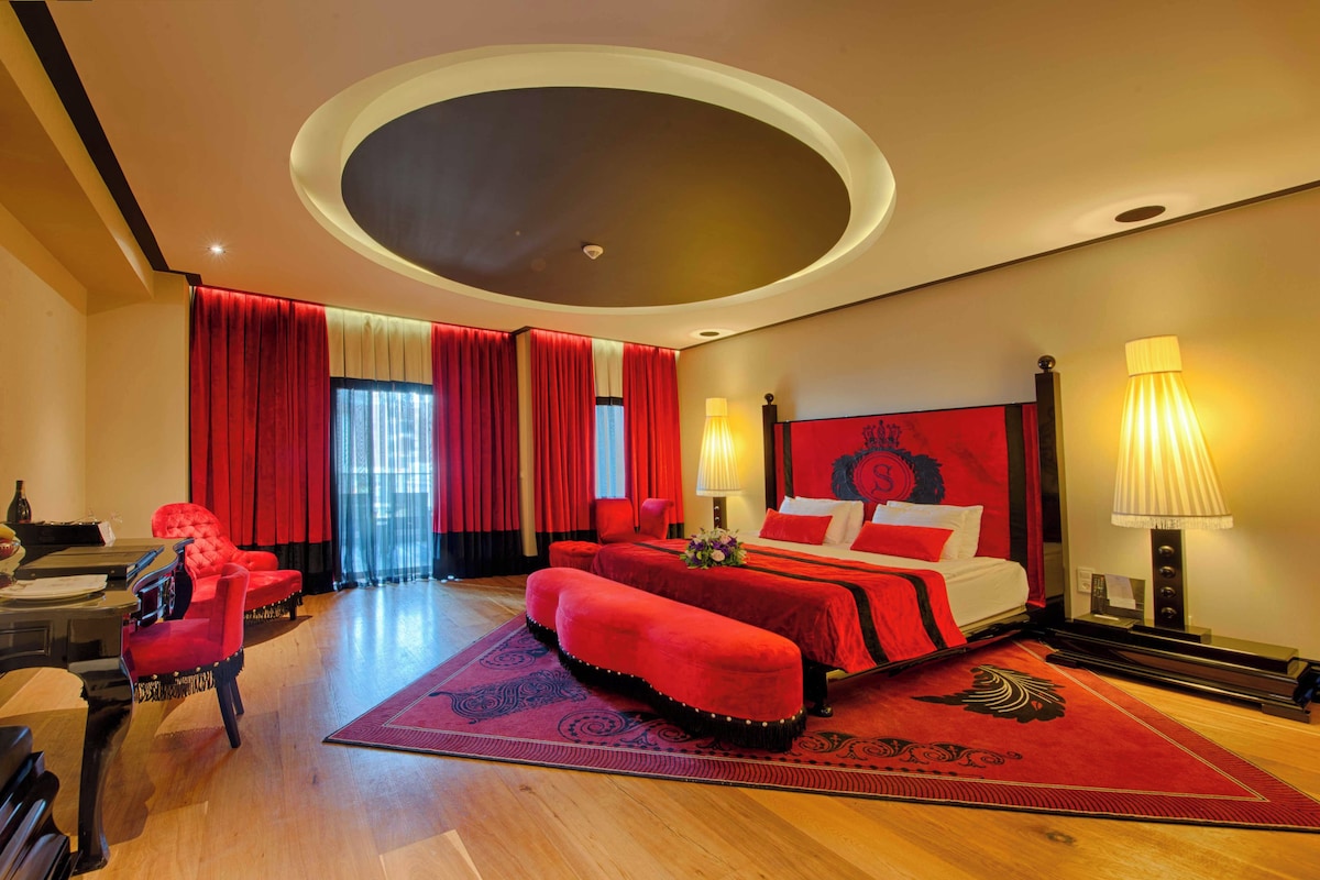 Une chambre d’hôtel avec un lit, un canapé rouge, un piano et une grande fenêtre avec des rideaux.