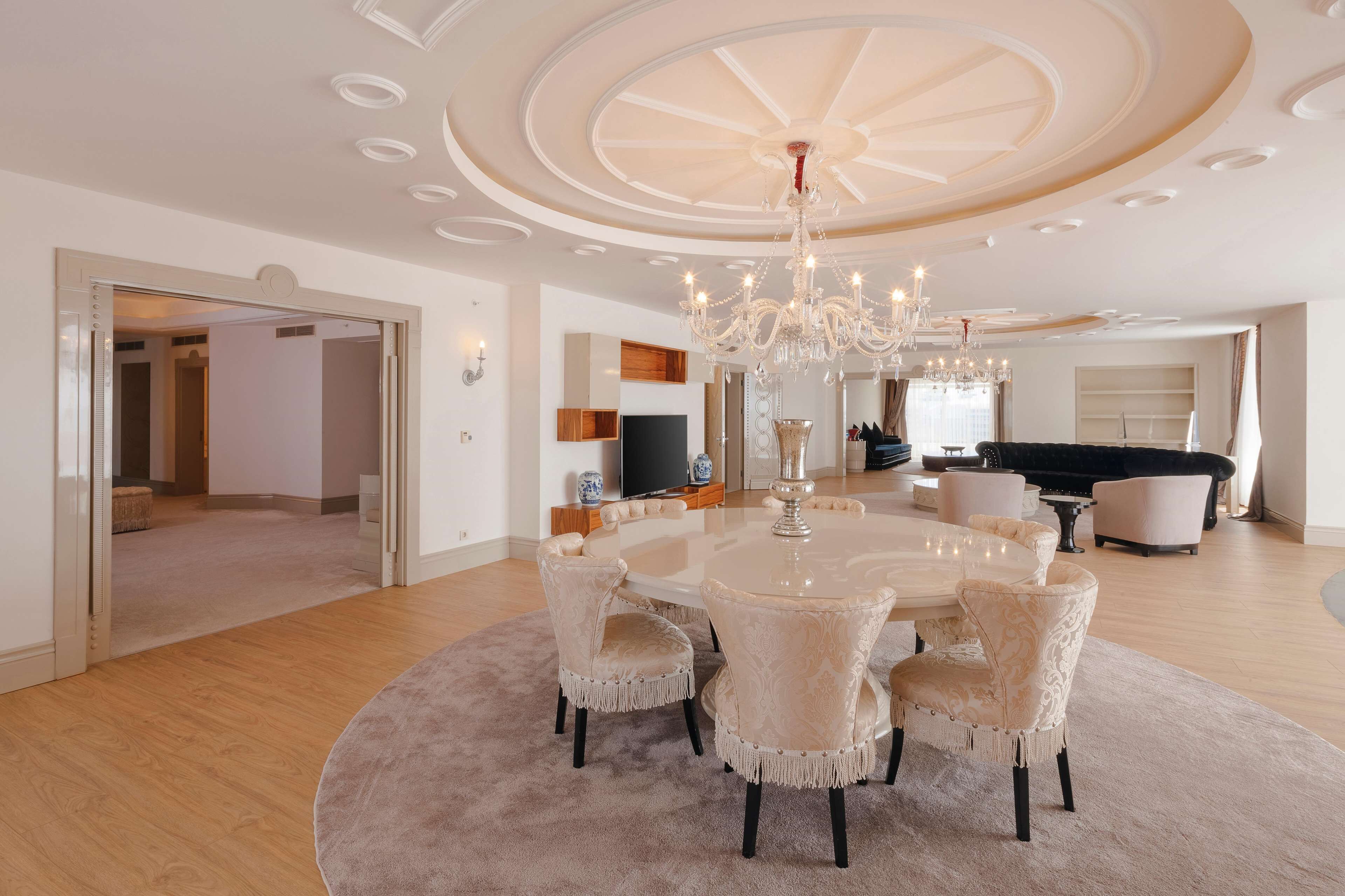A spacious dining area with a round table and six chairs, a chandelier, and a modern ceiling design.