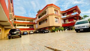 Un edificio de varios pisos con un patio empedrado, varios coches estacionados y una furgoneta blanca.