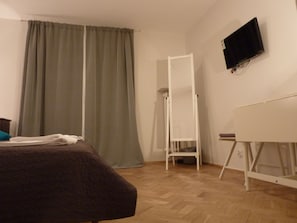 Une pièce comprenant un lit, un bureau debout, un téléviseur fixé au mur et une fenêtre avec des rideaux.