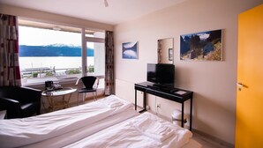 A hotel room with a bed, a desk with a TV, a chair, a small table with a coffee maker, and a window with a view of mountains and a lake. - Kringsjå Hotell (Sogndal)