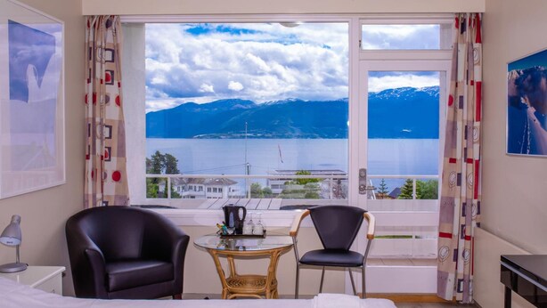 A hotel room with a bed, two chairs, a small table with a kettle, and a view of a lake and mountains. - Kringsjå Hotell (Sogndal)