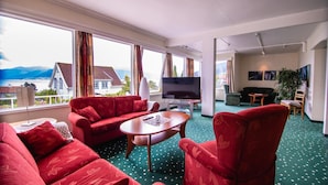 A spacious living room with red sofas, a wooden coffee table, and a flat-screen TV. Large windows offer a view of a house and mountains. - Kringsjå Hotell (Sogndal)