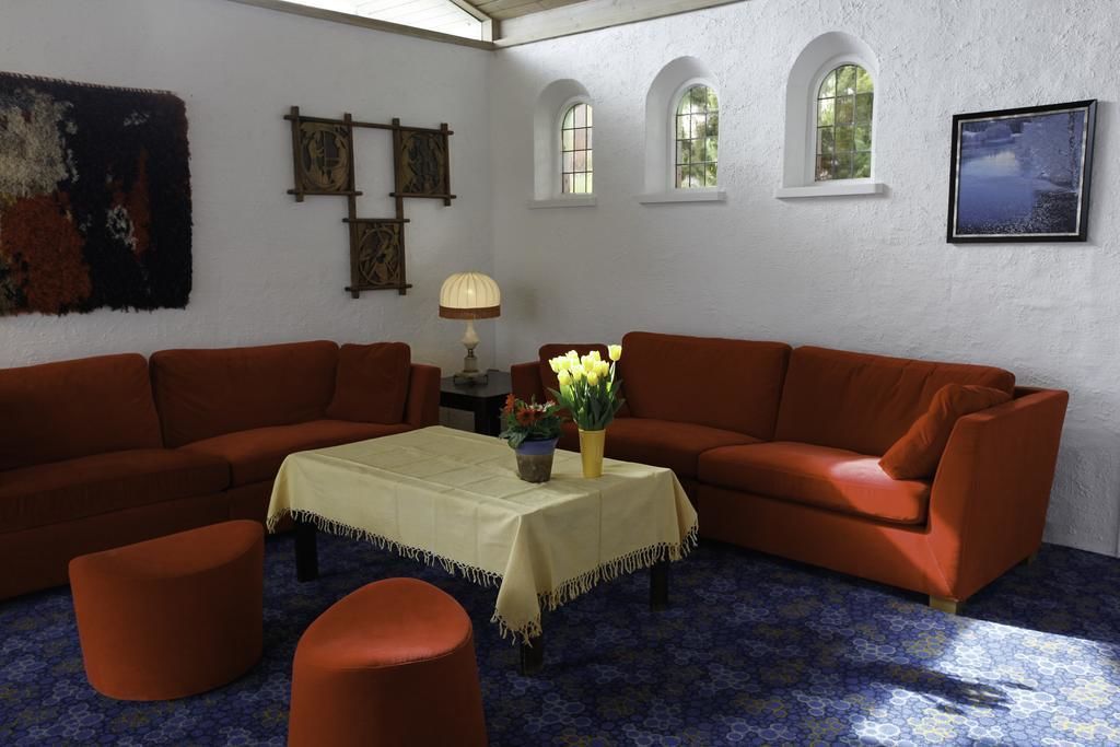 A room with two orange sofas, a small table with a yellow tablecloth, and a vase with flowers. There are three windows and a painting on the wall.
