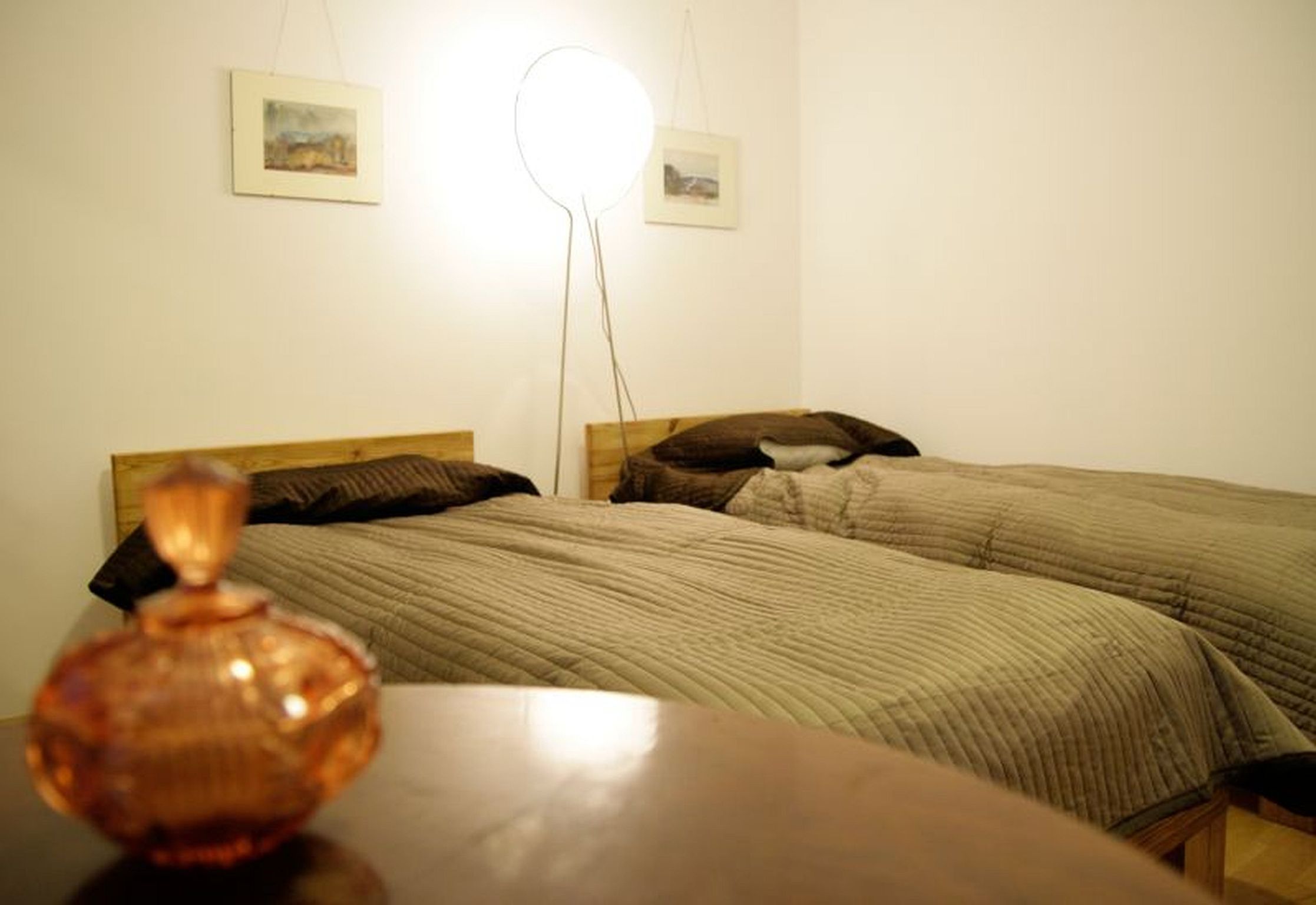 A room with two beds, a bedside table, and a wall-mounted light fixture.