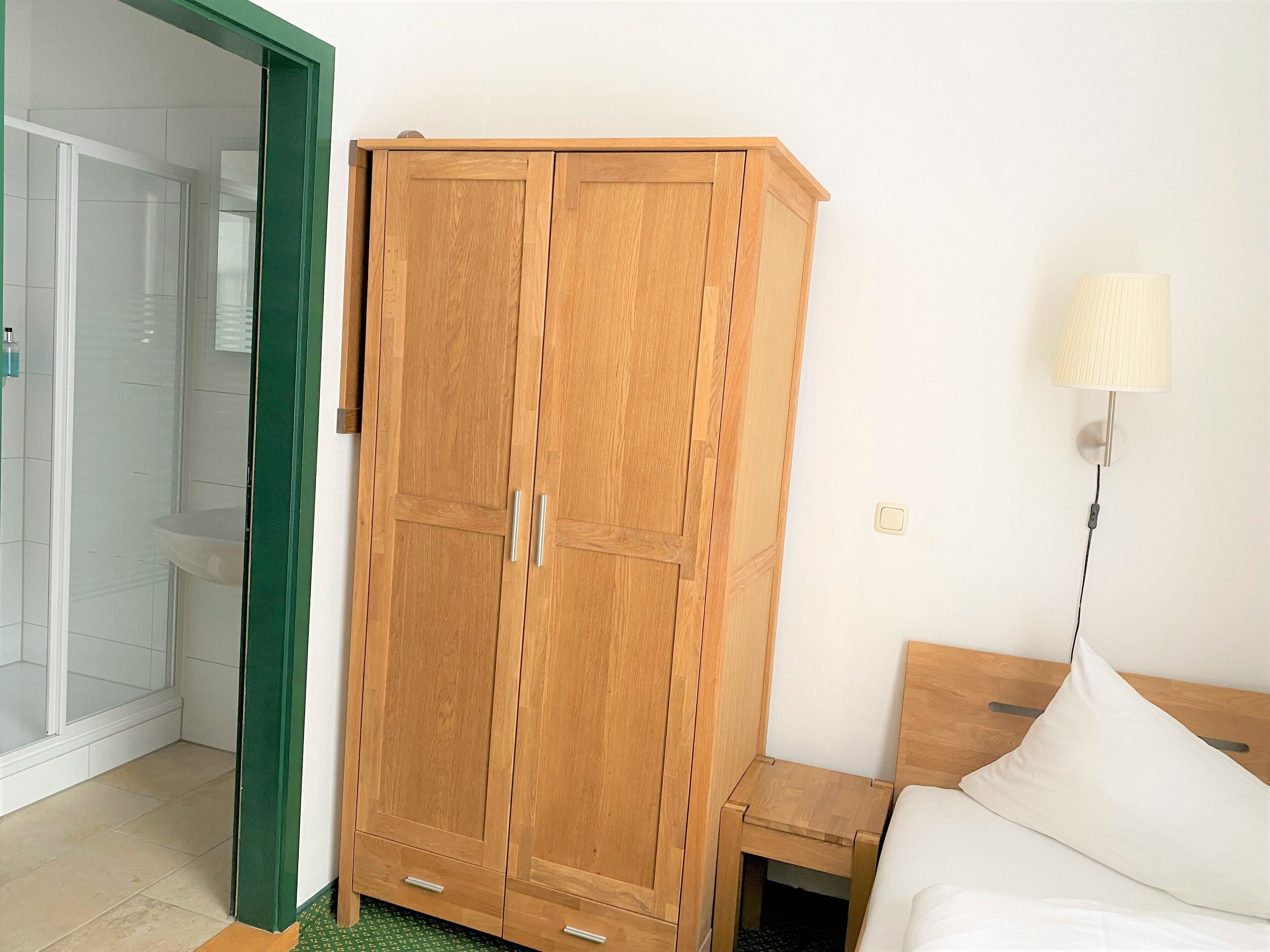 A bedroom with a wooden wardrobe, a bedside table, a lamp, and a green-framed door.