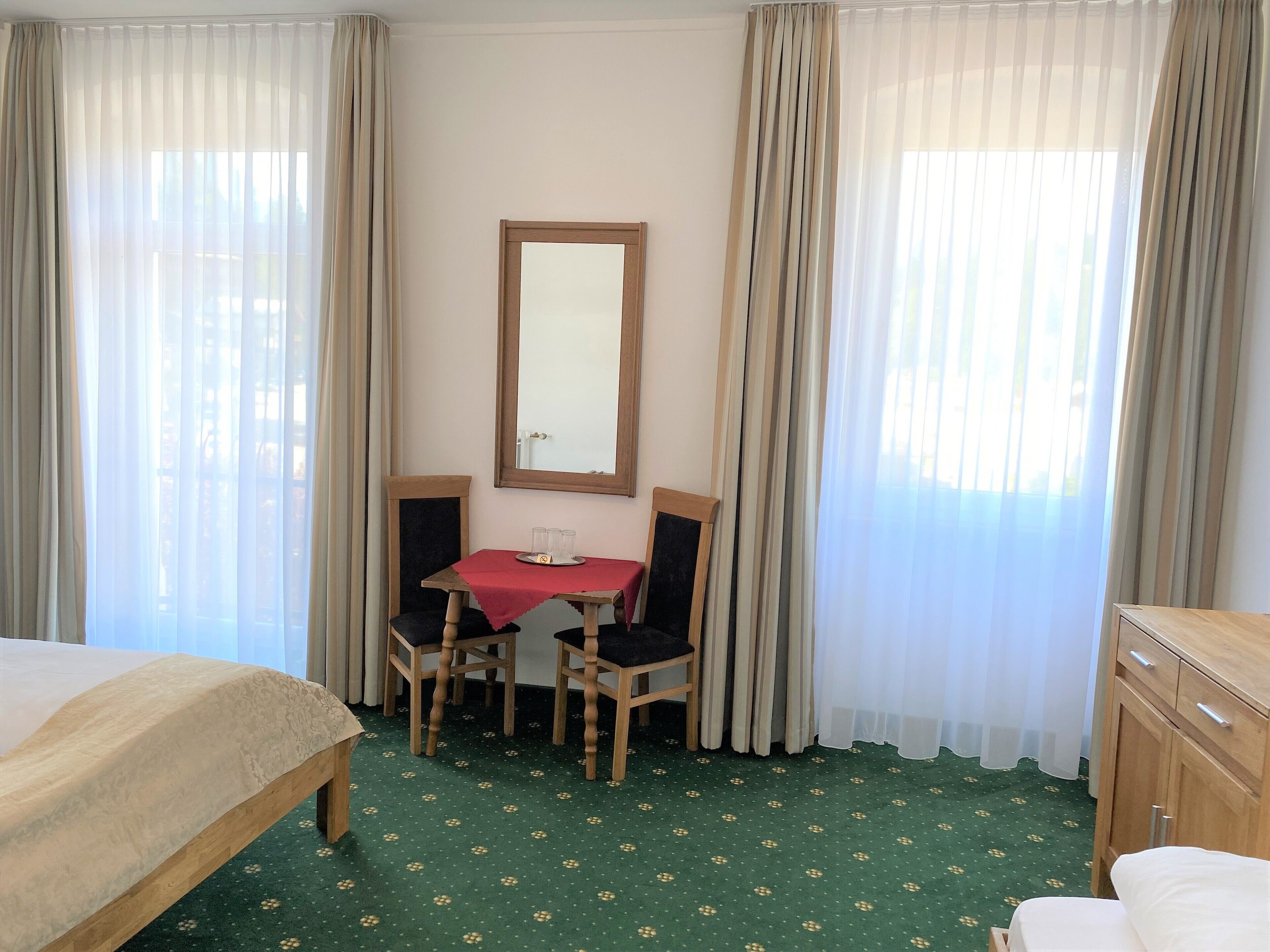 A hotel room with a bed, a desk with two chairs, a mirror, and a green patterned carpet.