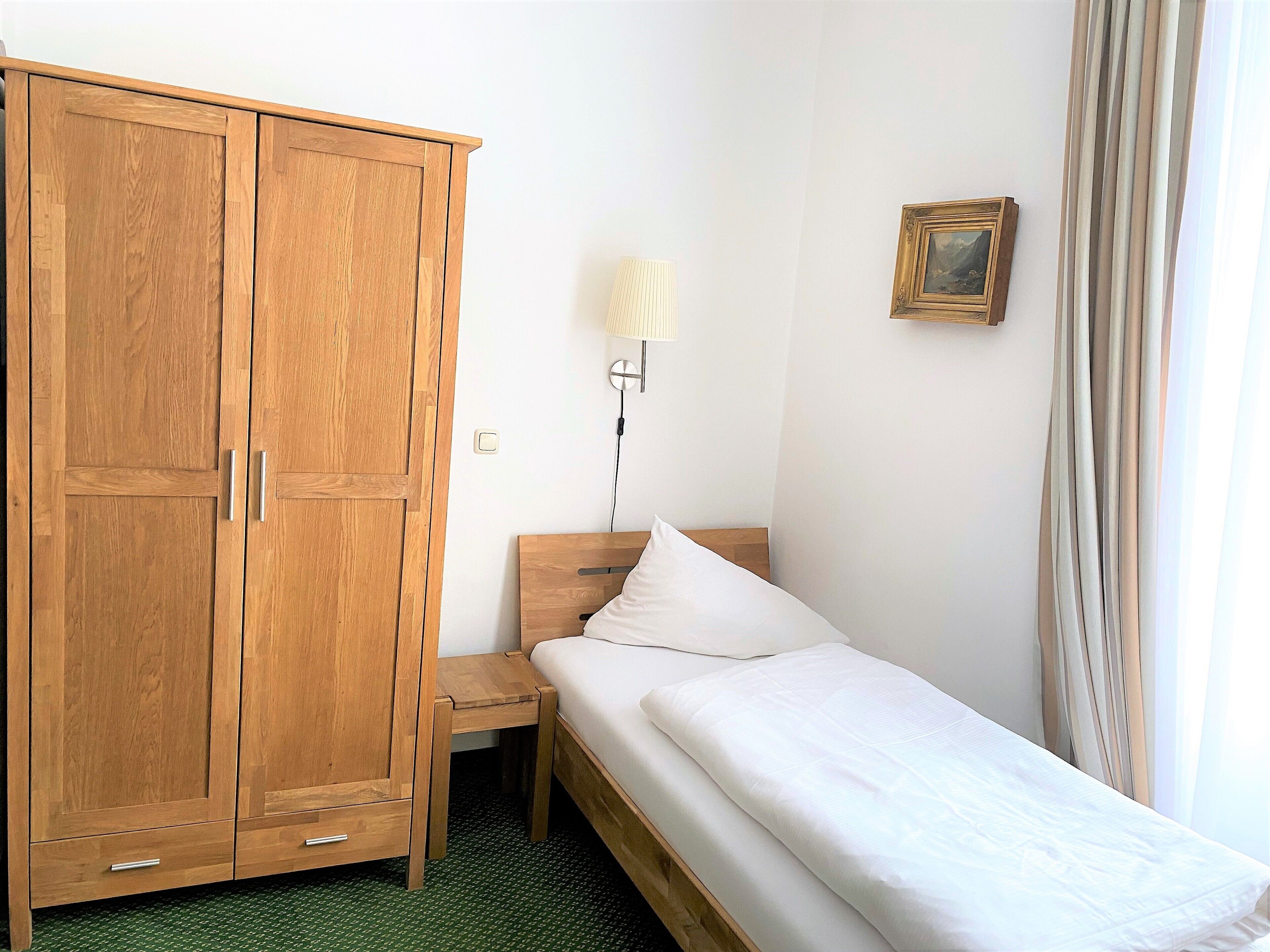 Ein Schlafzimmer mit Holzschrank, Nachttisch, Bett mit weißer Bettwäsche, Wandlampe und einem gerahmten Bild.