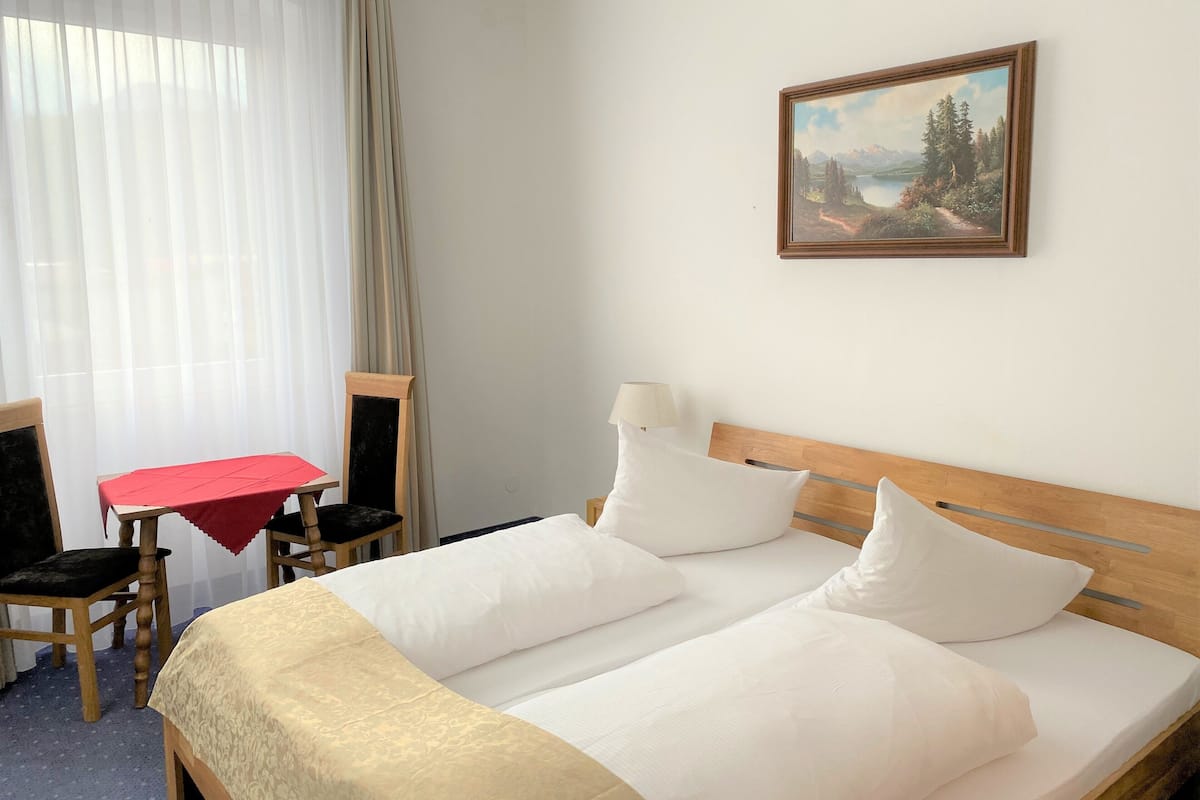 Habitación de hotel con cama doble, cabecera de madera, una mesita con mantel rojo, una silla y un cuadro en la pared.