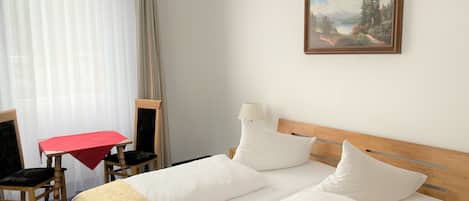 Habitación de hotel con cama doble, cabecera de madera, una mesita con mantel rojo, una silla y un cuadro en la pared.