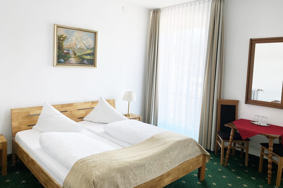 Habitación de hotel con cama de madera, un escritorio con mantel rojo, una silla, una lámpara, un cuadro en la pared y un ventanal con cortinas.