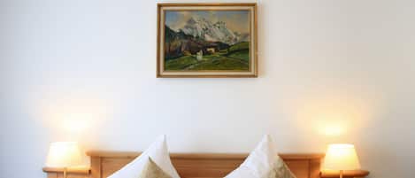 A hotel room with a bed, two bedside lamps, and a framed painting of a mountain landscape on the wall.