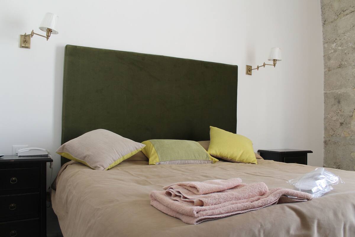 Una cama bien tendida con cabecera verde, dos mesitas de noche y una lámpara de pared.