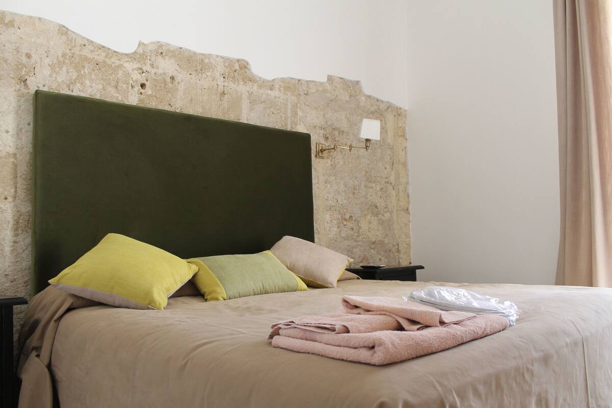 Una cama con cabecera verde, varios almohadones y toallas dobladas.