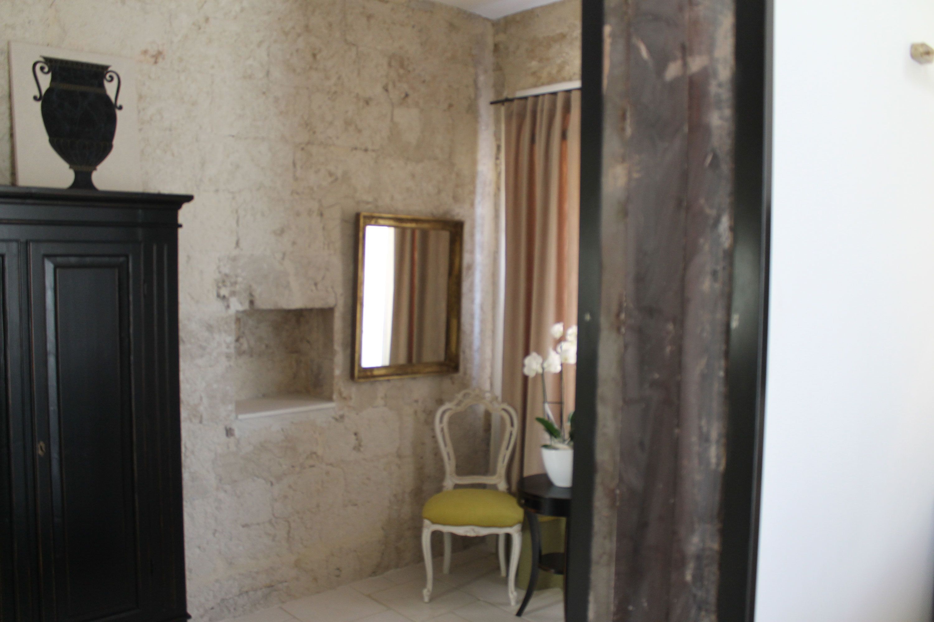 Une pièce avec un mur en pierre, une armoire en bois, une chaise avec un coussin, un miroir et une plante en pot.