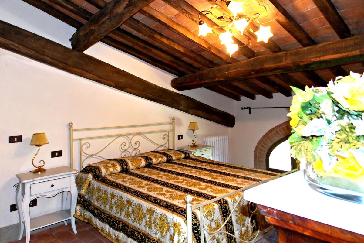 Quarto com cama, mesas de cabeceira, abajur, vaso de flores e vigas de madeira aparente.