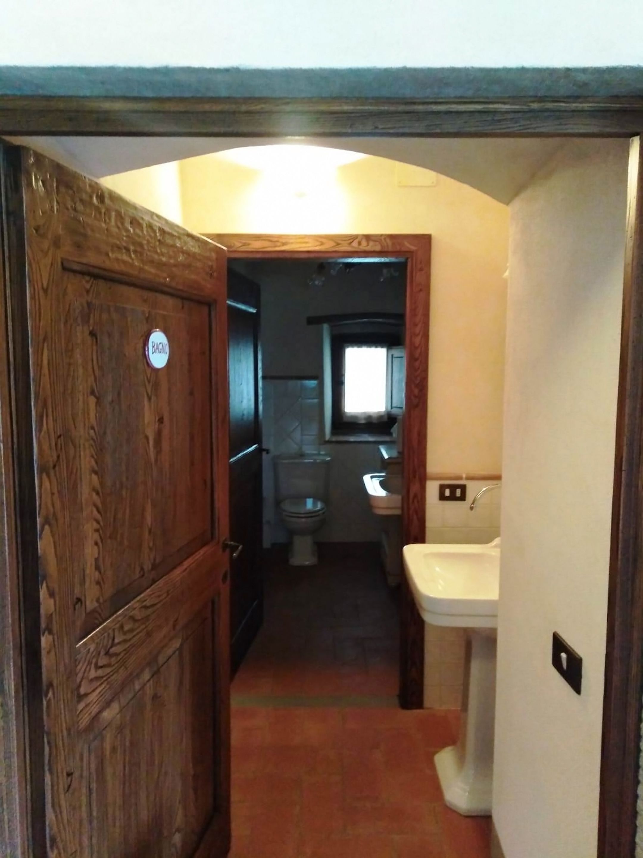 Un couloir étroit mène à une salle de bain équipée de toilettes, d’un lavabo et d’une porte en bois sur laquelle est apposée une pancarte.