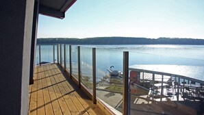 Um deck de madeira com guarda-corpos de vidro, vista para um lago, um barco e uma margem florestada ao longe.