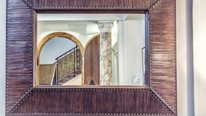 A mirror with a dark wooden frame and metallic studs, reflecting a staircase with a wooden handrail and a marble column.