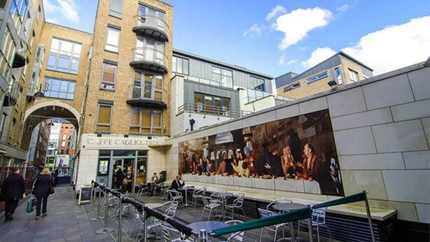 Un área de café al aire libre con mesas y sillas, un mural en la pared y un edificio con un letrero que dice "CAFFE CAGLIOSTRO".