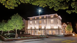 A multi-story hotel with a balcony on the second floor, a well-lit entrance, and a parking area with cars.