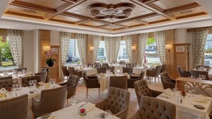 A restaurant with multiple dining tables and chairs, large windows with curtains, and a wooden ceiling with intricate designs.