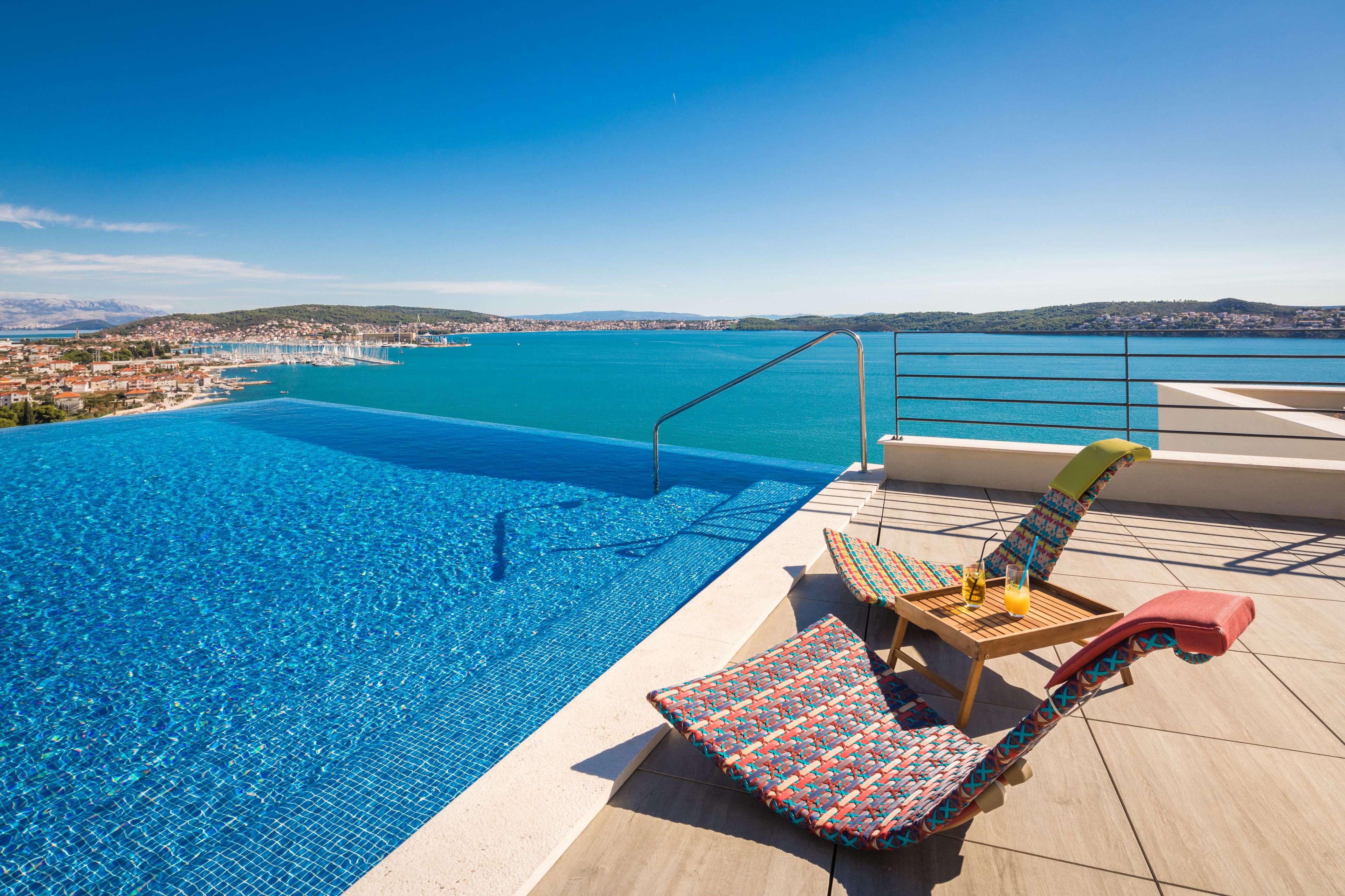 A swimming pool with a view of a coastal town, a lounge chair, and a table with a drink.