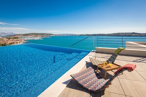 A swimming pool with a view of a coastal town, a lounge chair, and a table with a drink. - Hotel Ola - Adults Only (Seget)