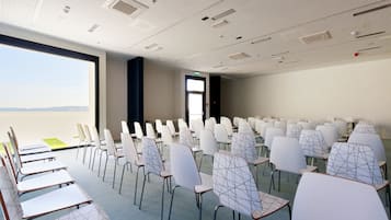 Une salle de conférence avec des rangées de chaises et de tables blanches, une grande fenêtre offrant une vue dégagée et un plafond doté d’un éclairage encastré et d’un système de ventilation.