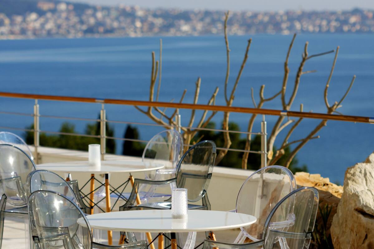 A table with clear chairs and a white tablecloth, overlooking a coastal cityscape with a view of the sea.