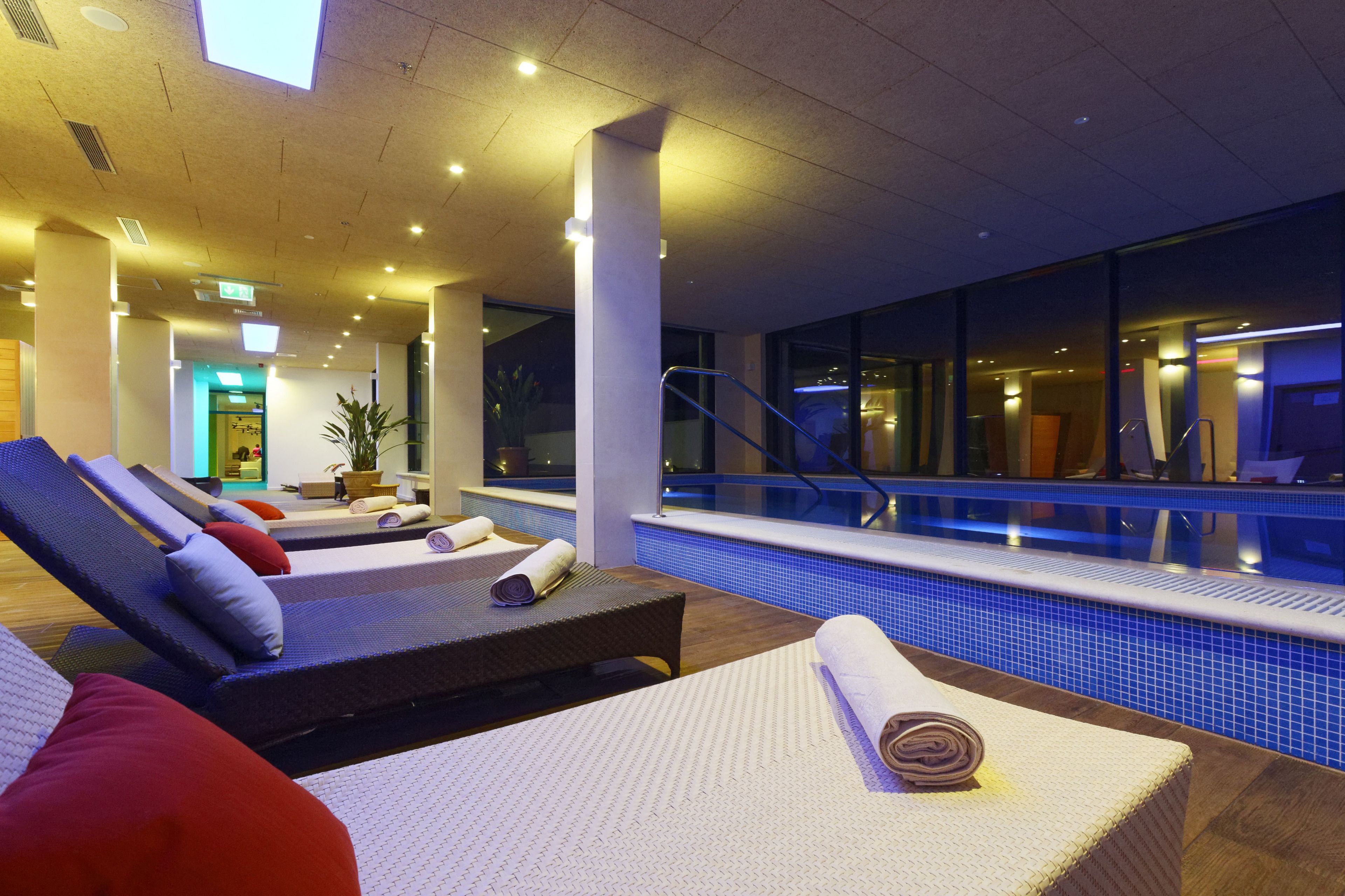 A poolside area with lounge chairs and a small table, a pool with a blue tiled edge, and a staircase leading to an upper level.