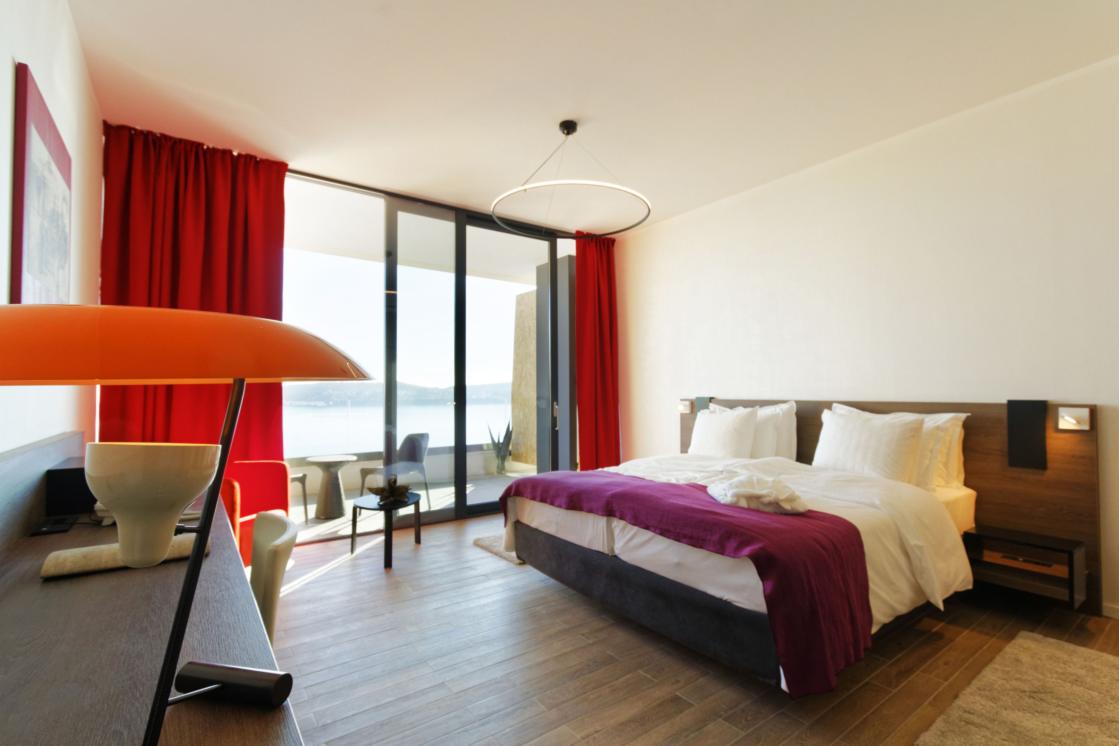 A modern hotel room with a large bed, a desk with a chair, a view of the outdoors, and a large window with red curtains.