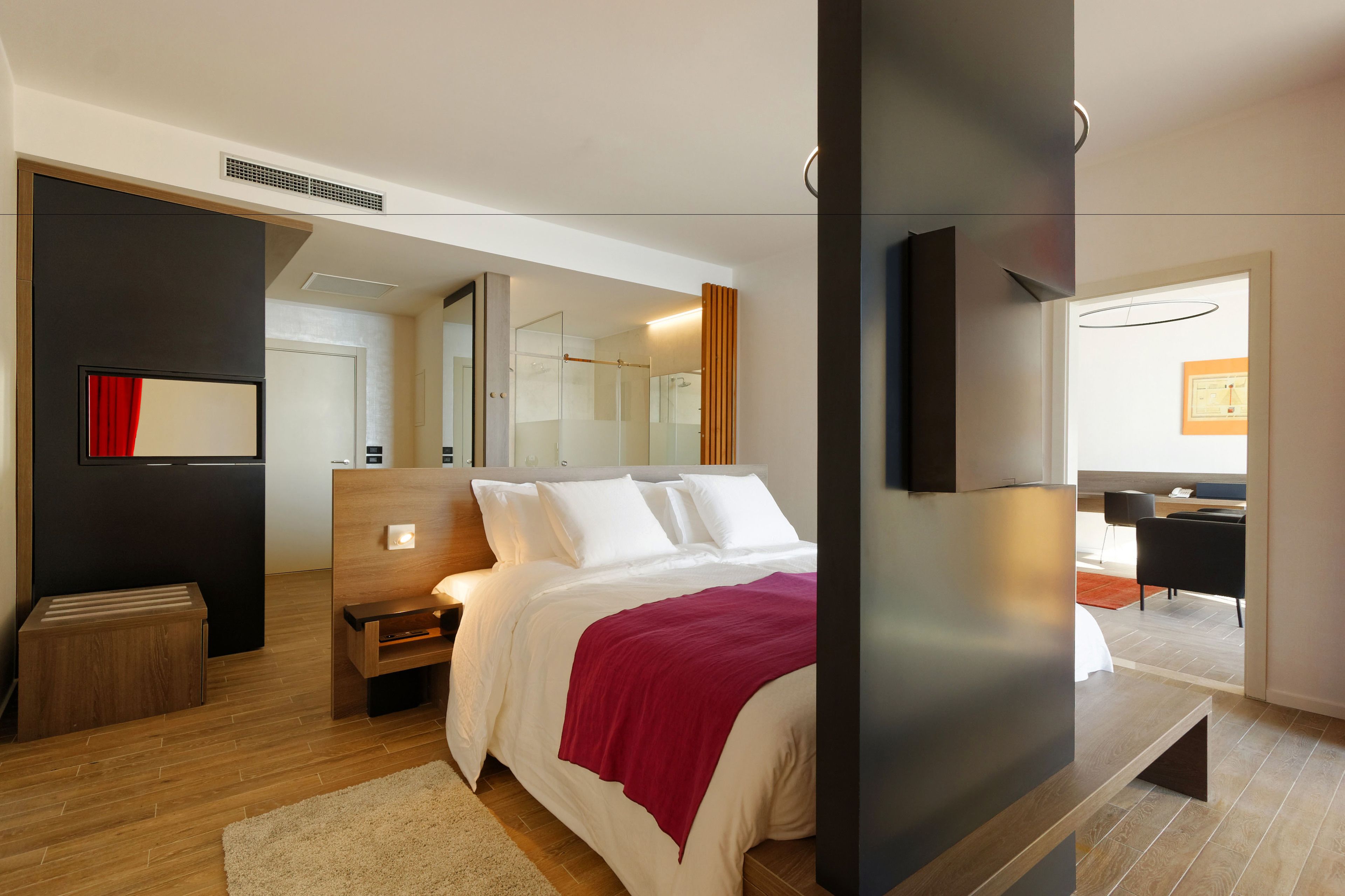 Quarto de hotel moderno com uma cama grande, uma televisão fixada na parede, uma mesinha de cabeceira de madeira e um espelho.