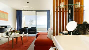 A modern hotel room with a large bed, a seating area with a table and chairs, a small balcony, and a bathroom with a mirror and toiletries.
