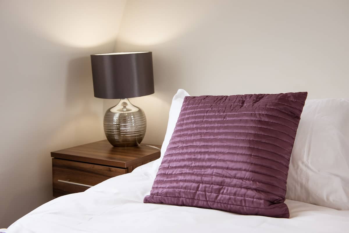A neatly made bed with a white pillow and a purple pillow, a wooden nightstand with a lamp, and a drawer.