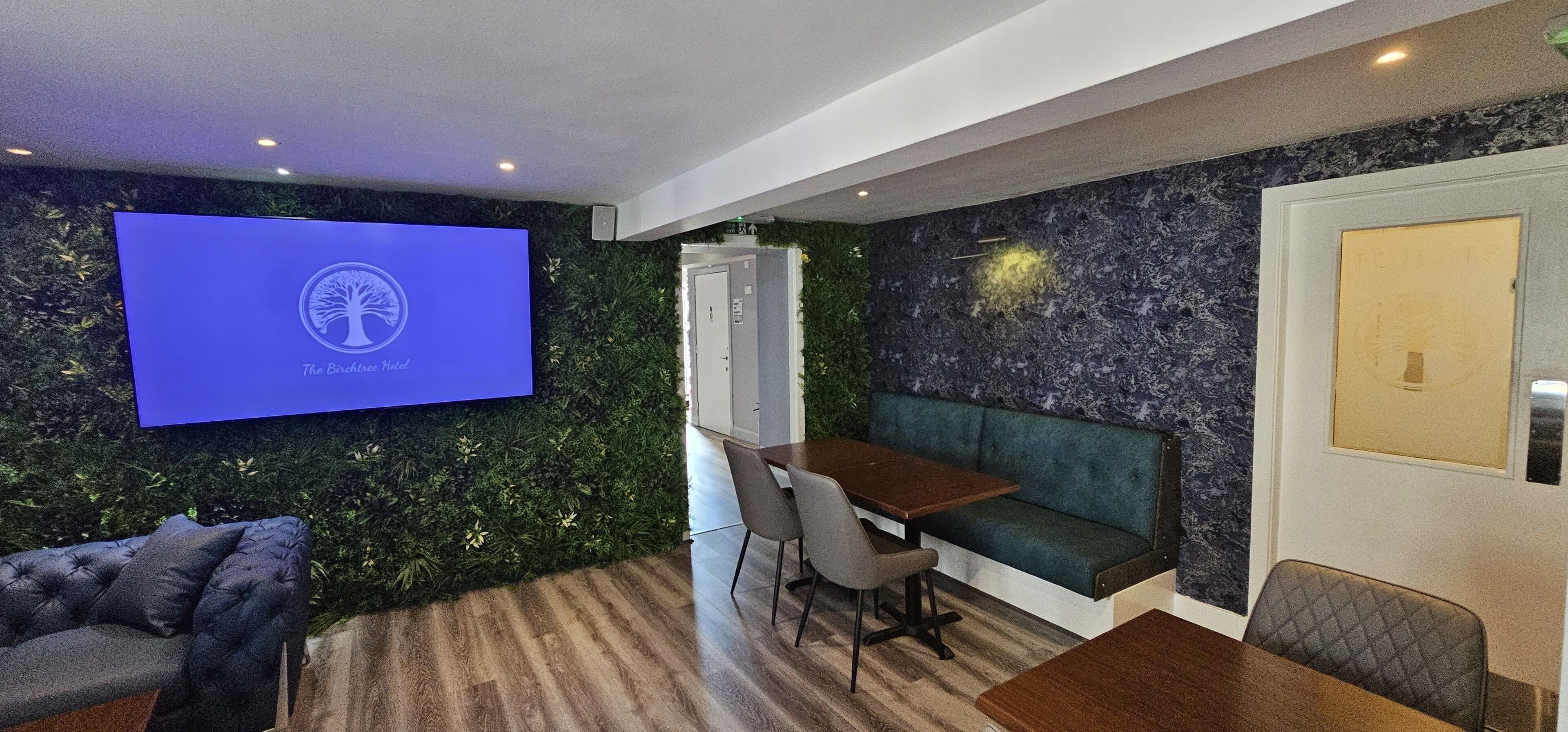 Een moderne hotelkamer met een groene wand, een flatscreen televisie waarop een logo staat, een houten eettafel, een bank en een lange zitbank.