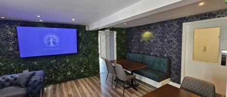 Een moderne hotelkamer met een groene wand, een flatscreen televisie waarop een logo staat, een houten eettafel, een bank en een lange zitbank.