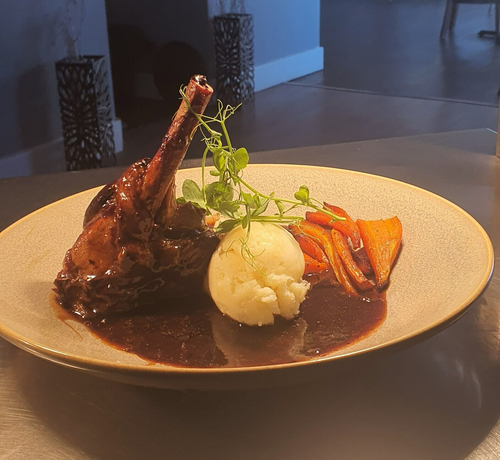 A plate with a lamb shank, mashed potatoes, and roasted carrots.