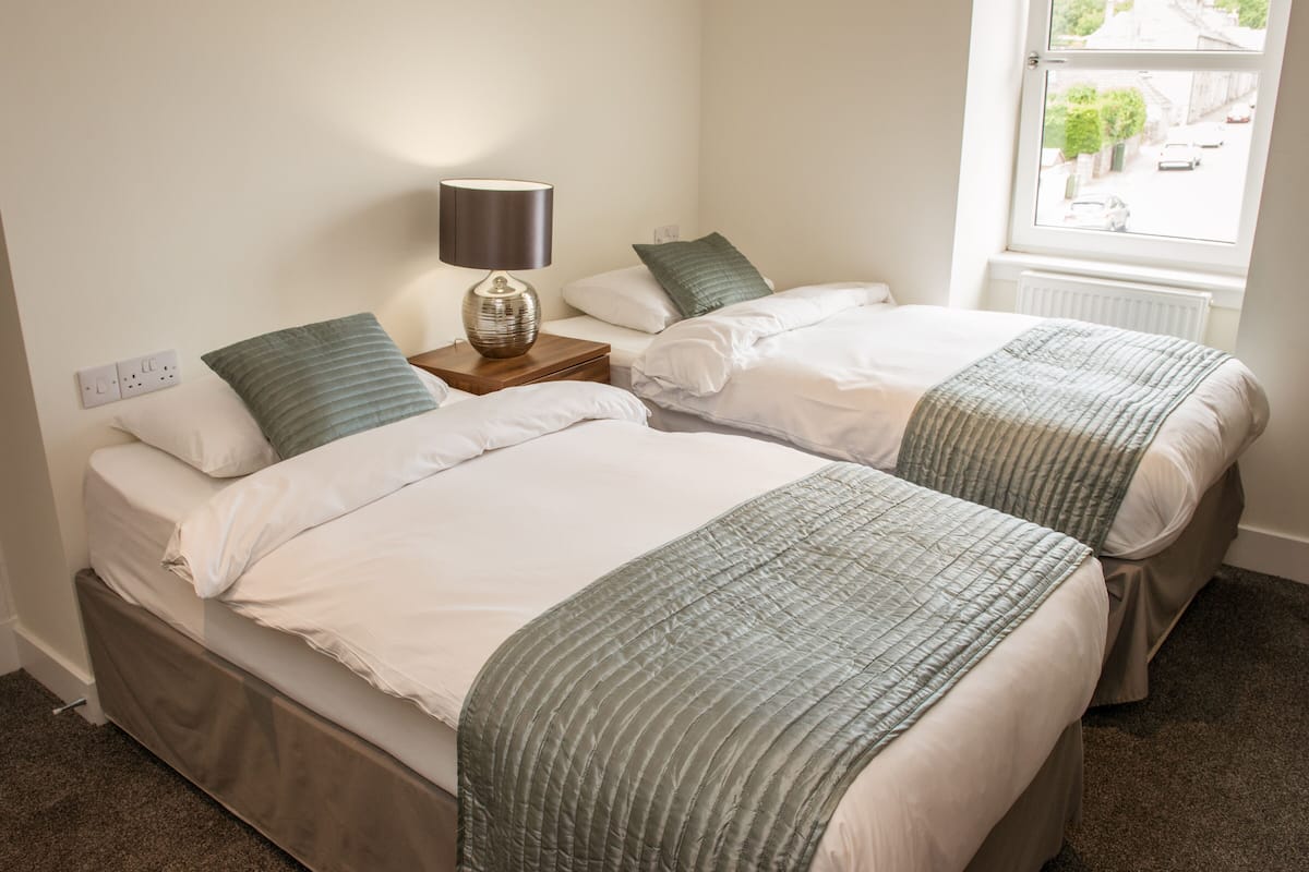 A bedroom with two beds, a nightstand with a lamp, a window with a view of the street, and a radiator.