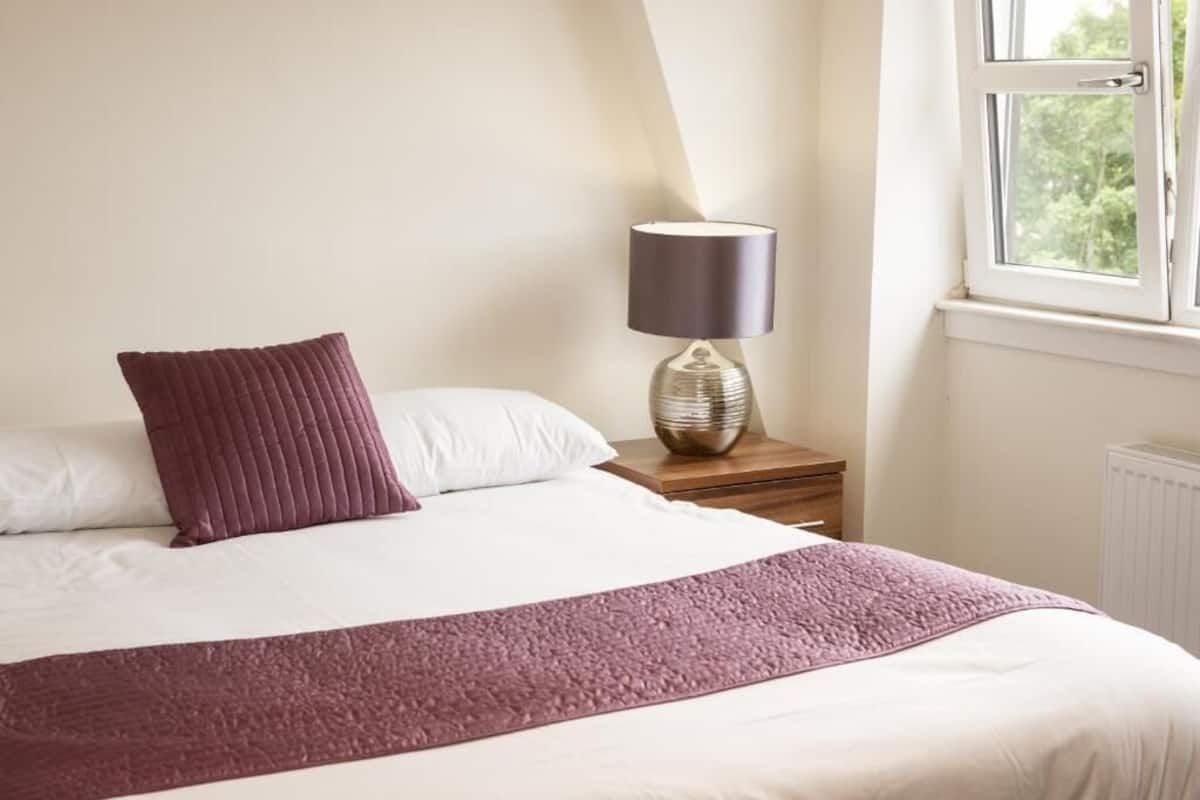 A neatly made bed with a white and purple blanket, a dark lamp on a wooden nightstand, and a window with a view of greenery.
