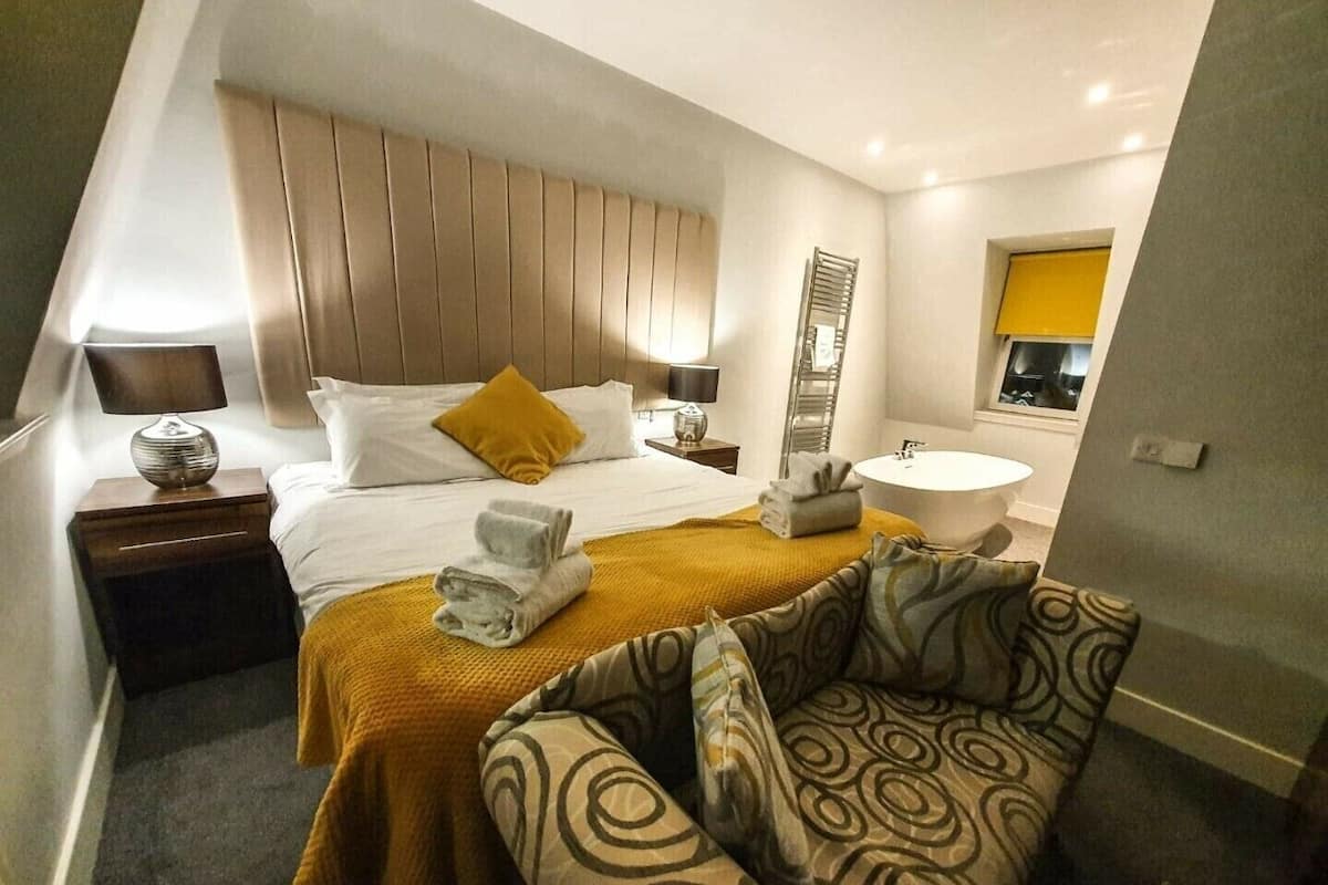 A modern hotel room with a bed, a sofa, a bedside table with a lamp, a mirror, a sink, and a towel rack.