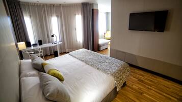 Habitación de hotel con una cama grande, un escritorio con silla, un televisor en la pared y una ventana con cortinas.