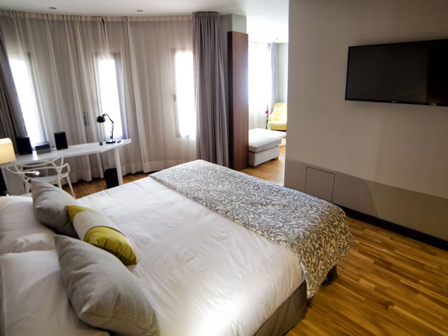 Habitación de hotel con una cama grande, un escritorio con silla, un televisor en la pared y una ventana con cortinas.