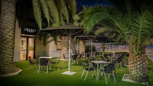 Un espace extérieur aménagé avec des tables et des chaises, des palmiers et un bâtiment sur lequel on peut lire l’inscription « GASSET ».