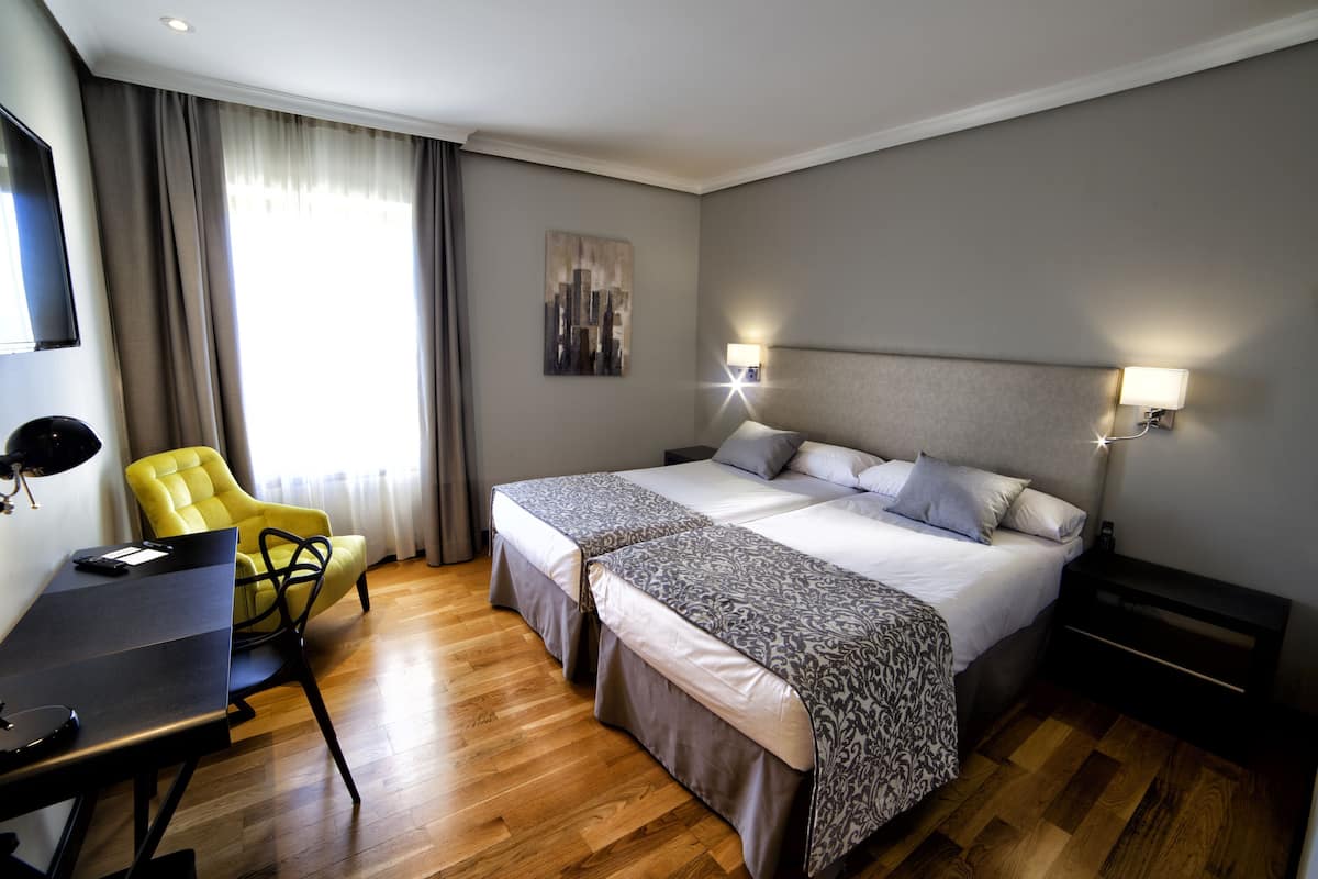 A hotel room with a double bed, a desk, a chair, and a cityscape painting.